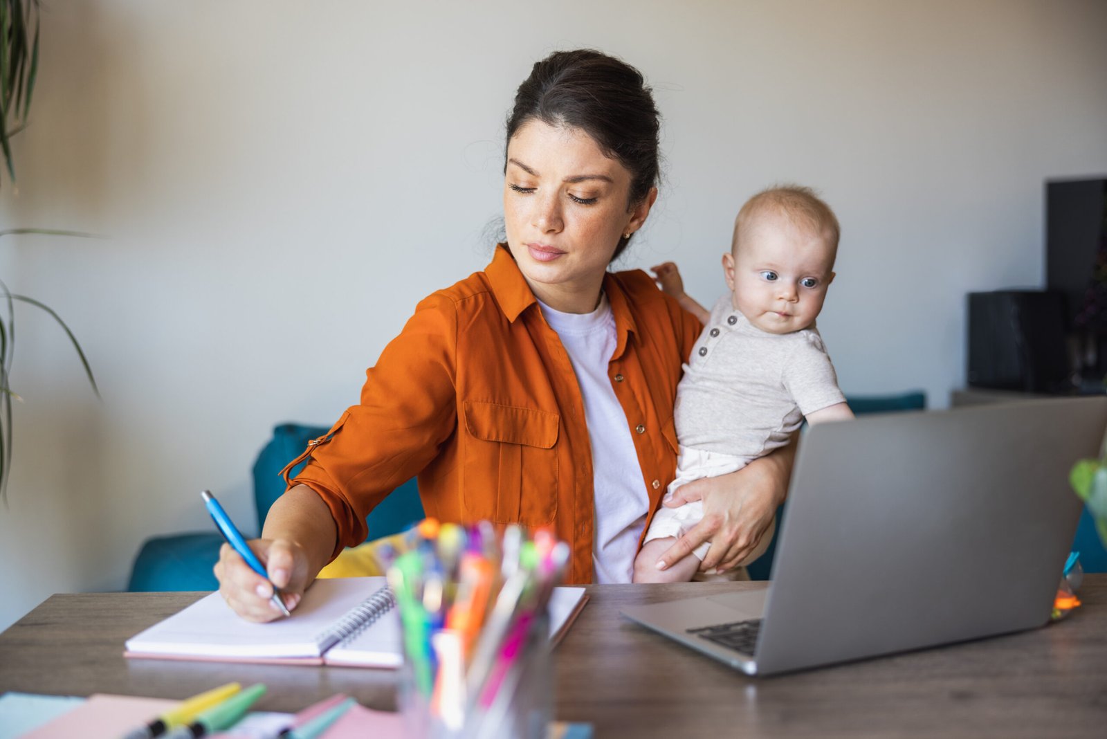 Busy mother writing in notes while babysitting her daughter and working at home.