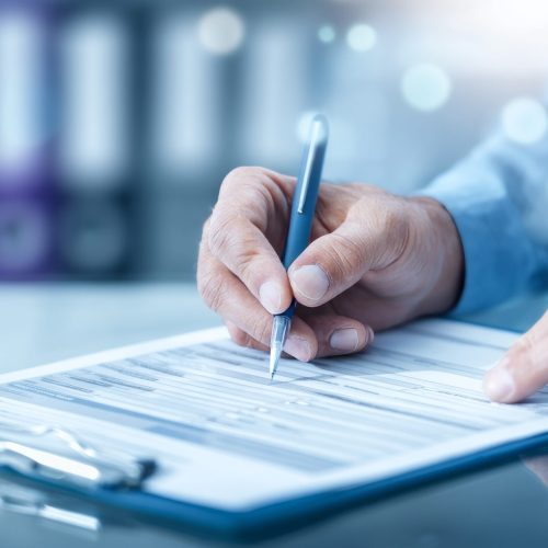 Closeup image of hands filling out patient intake forms while sitting at a table, conveying the administrative aspect of healthcare services and documentation.